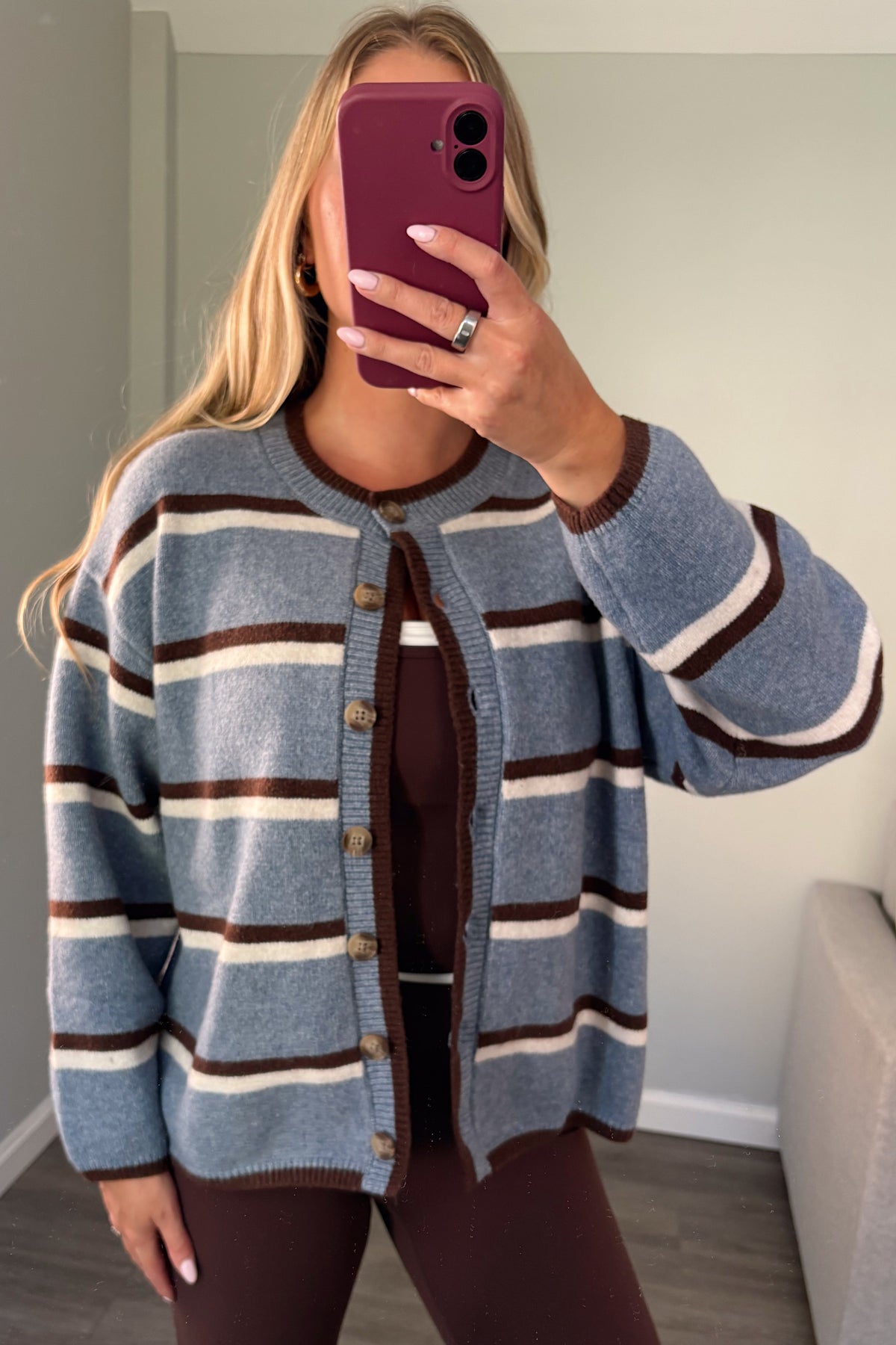 A woman takes a mirror selfie wearing a blue, brown, and cream striped knit cardigan over a dark brown top and matching brown leggings in a home setting.