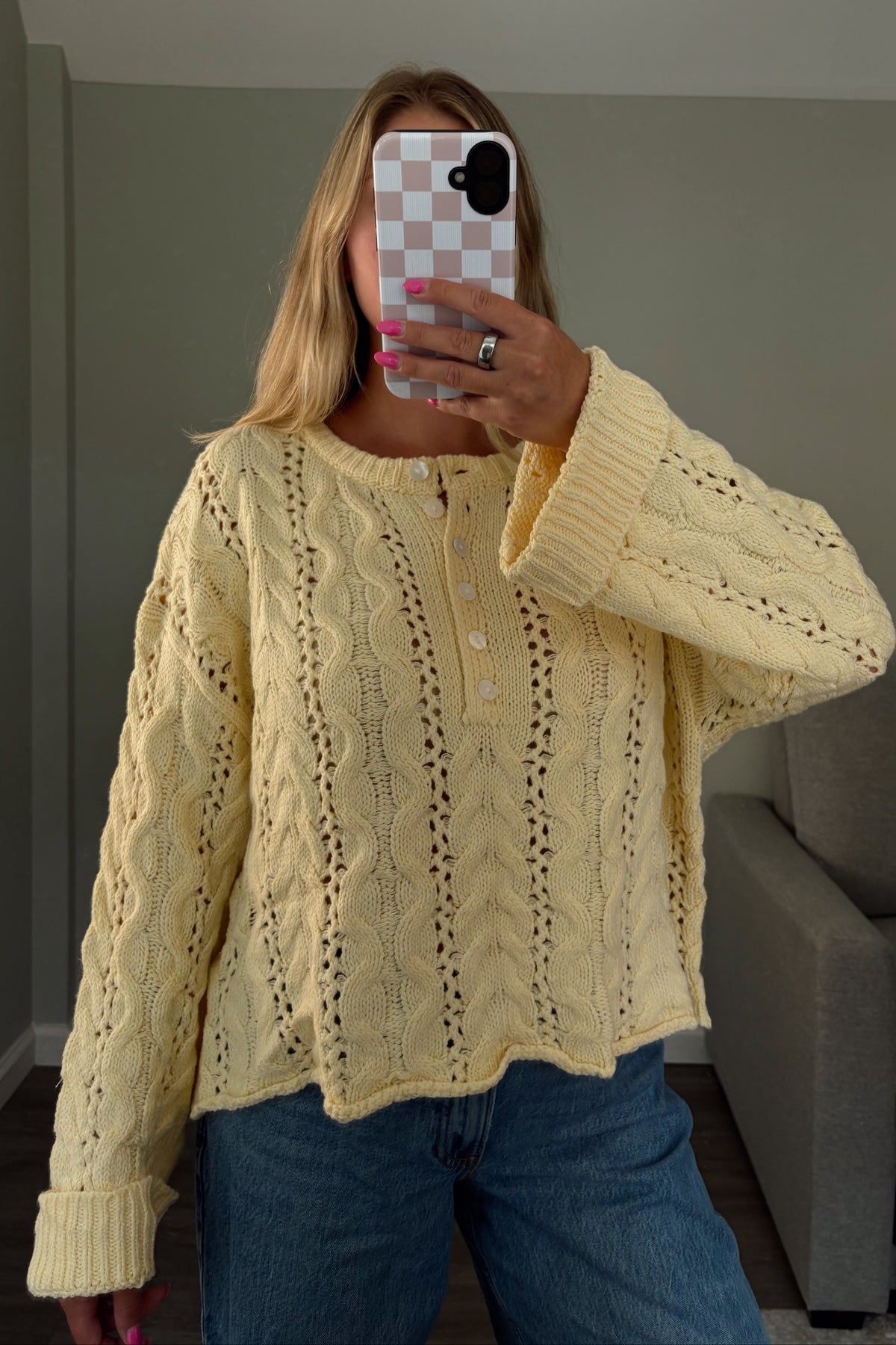 A woman takes a mirror selfie wearing a pale yellow cable-knit button-up sweater and blue jeans in a home setting with a gray wall and sofa in the background.
