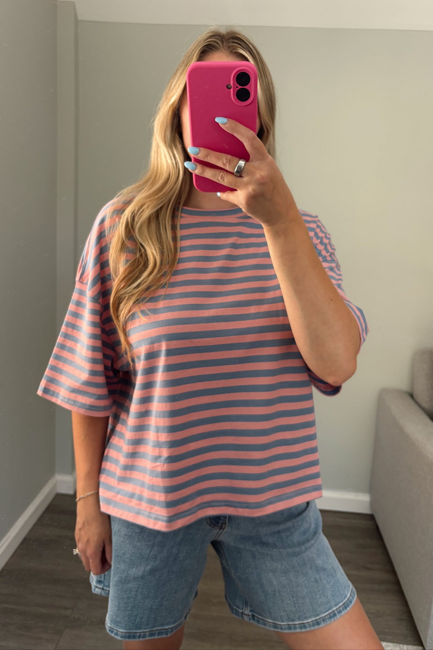 A woman takes a mirror selfie wearing a pink and light blue horizontally striped short-sleeve t-shirt and denim shorts in a home setting.