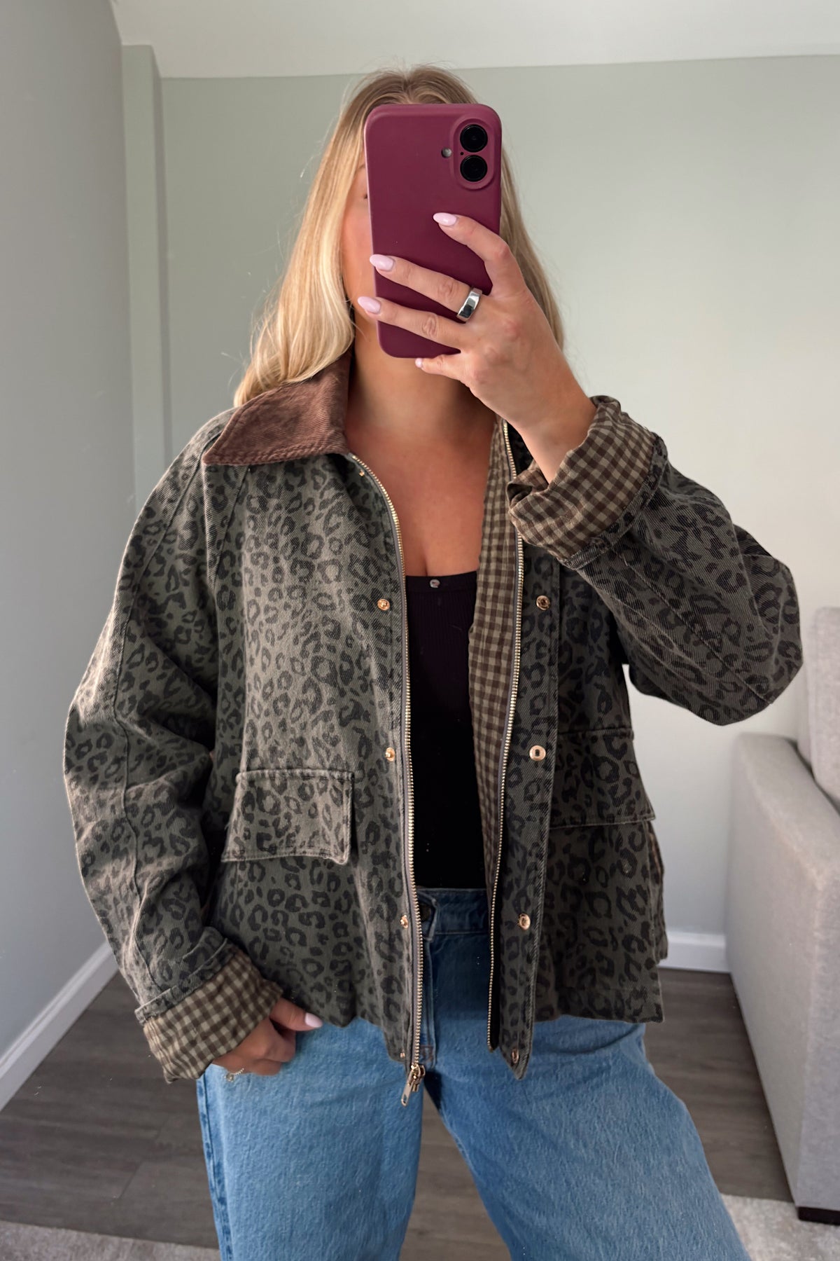A woman takes a mirror selfie wearing a brown and black leopard print jacket with a brown gingham lining and collar, paired with blue jeans and a black top.