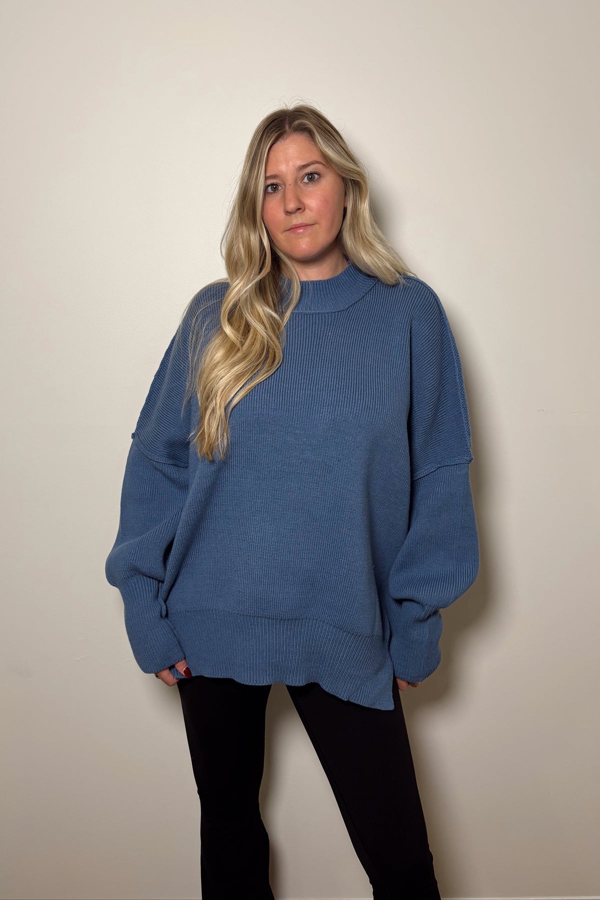Essential dusty blue oversized mock neck sweater worn with black leggings, showing dropped shoulders and a long ribbed hem.


