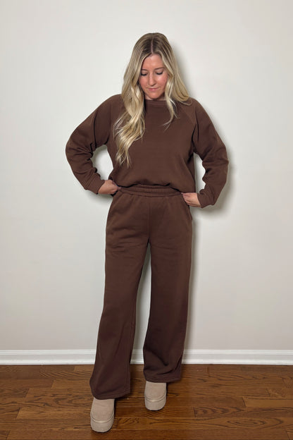 Woman wearing a brown sweat set/lounge set standing against a plain wall.