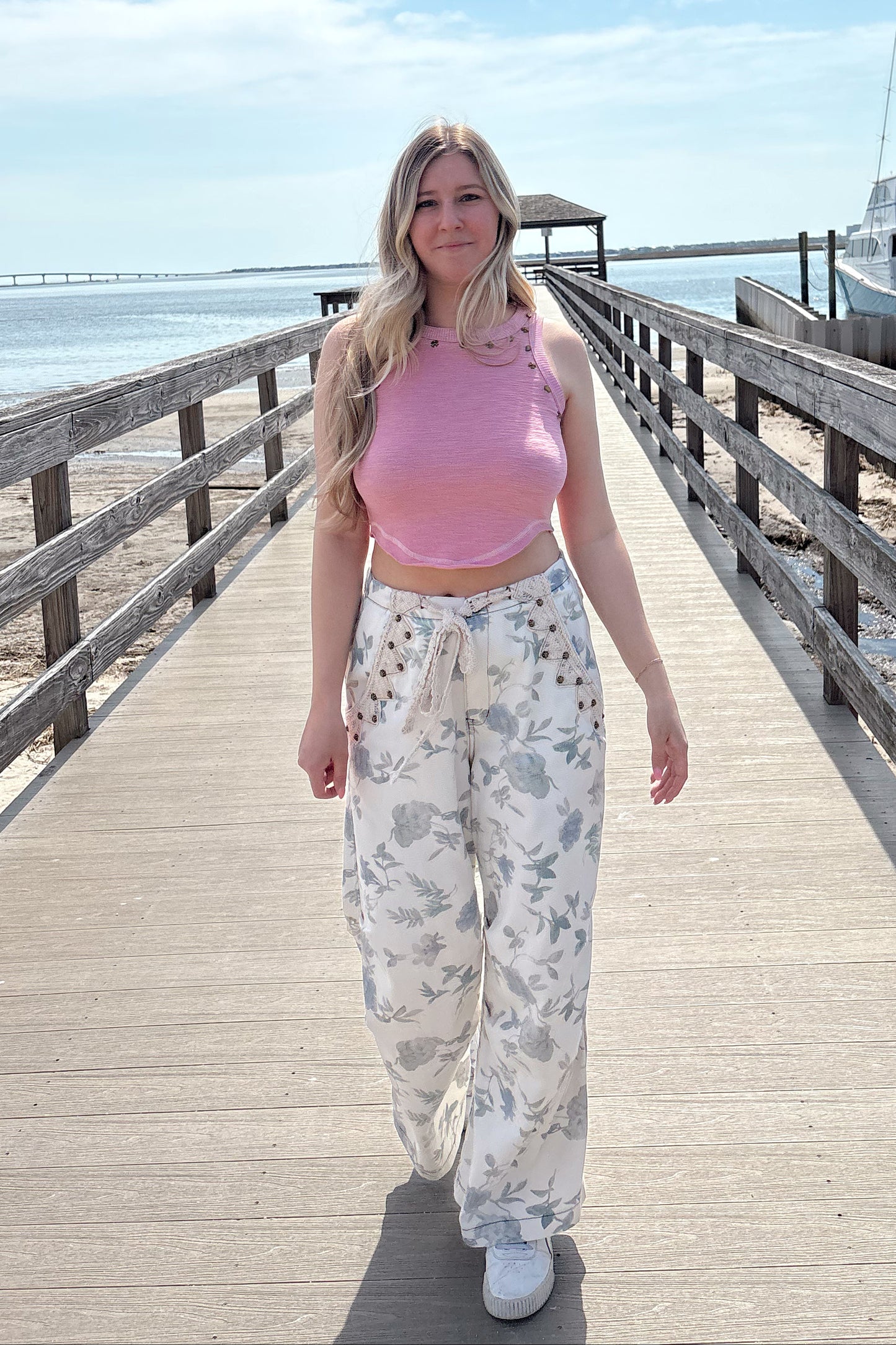 Woman in a pink top and white floral pants standing on a wooden pier with a clear blue sky.