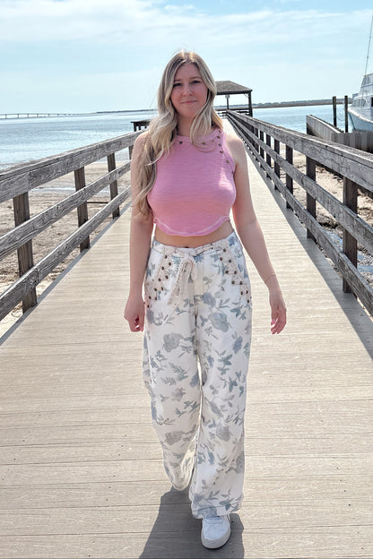 Woman in a pink top and white floral pants standing on a wooden pier with a clear blue sky.
