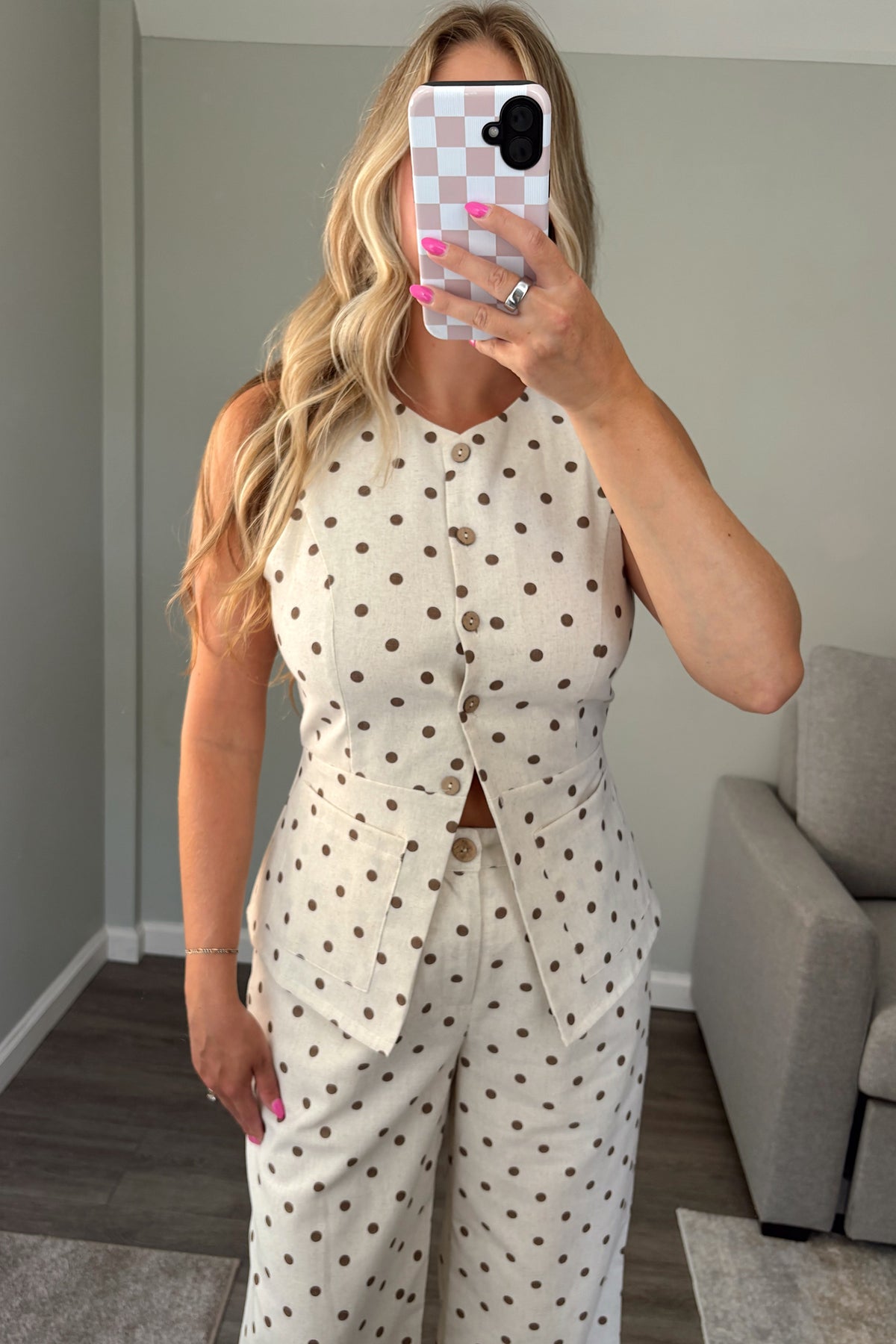 A woman takes a mirror selfie wearing a sleeveless cream top with brown polka dots and a button-front waist detail, in a modern room with a gray sofa and wood floor.
