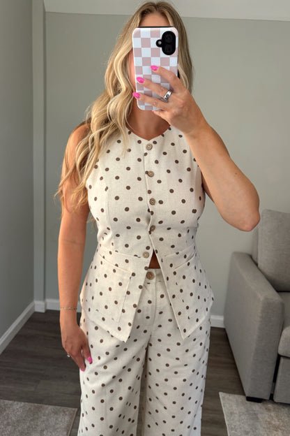 A woman takes a mirror selfie wearing a sleeveless cream top with brown polka dots and a button-front waist detail, in a modern room with a gray sofa and wood floor.