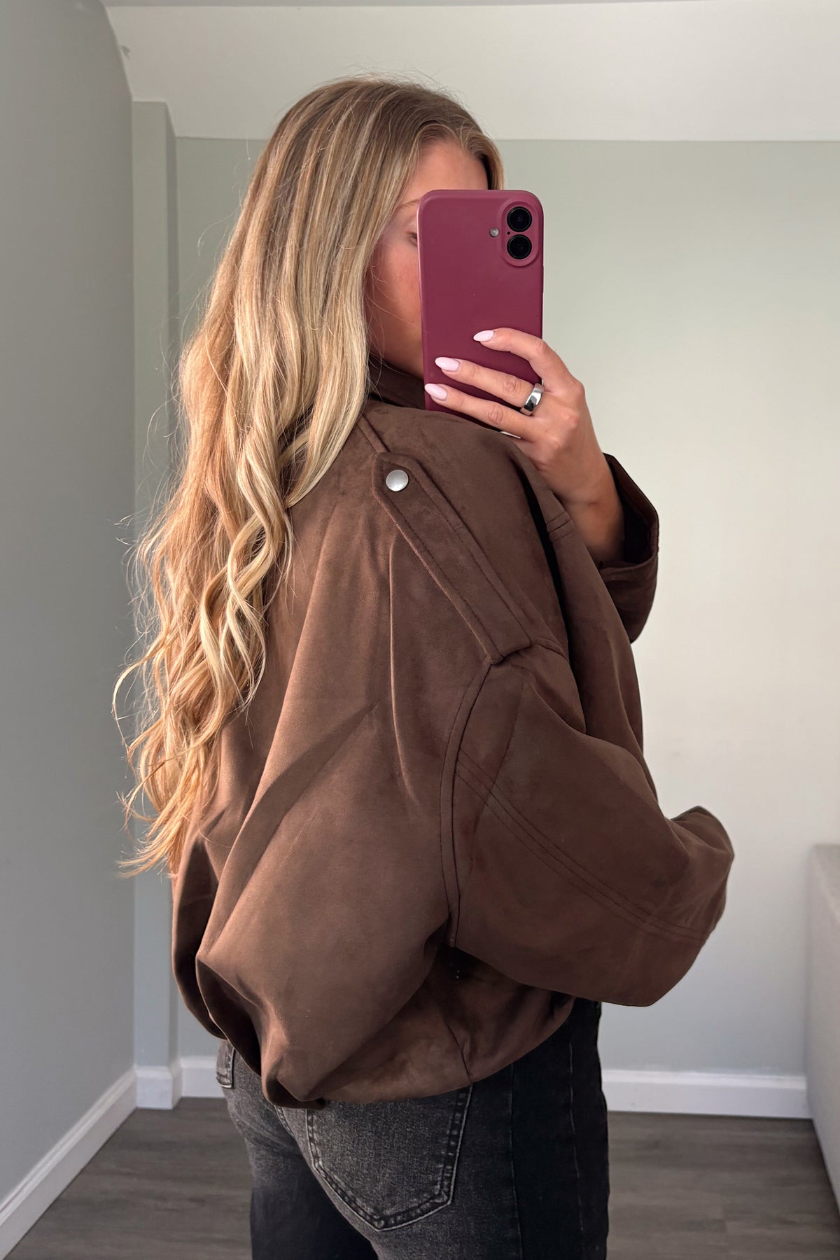 A woman takes a mirror selfie wearing an oversized brown jacket and dark gray jeans in a room with light gray walls.