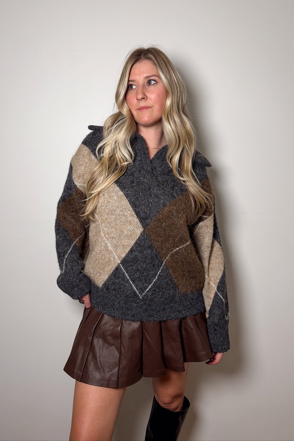 Blake charcoal, brown, and beige argyle collared sweater worn untucked over a brown pleated mini skirt with tall black boots.

