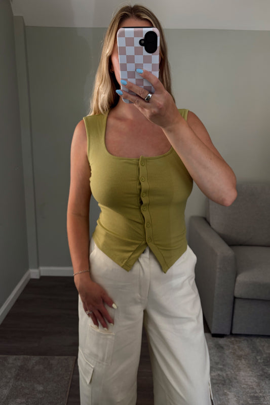 Woman taking a mirror selfie wearing a green top and white pants in a room with a gray couch.