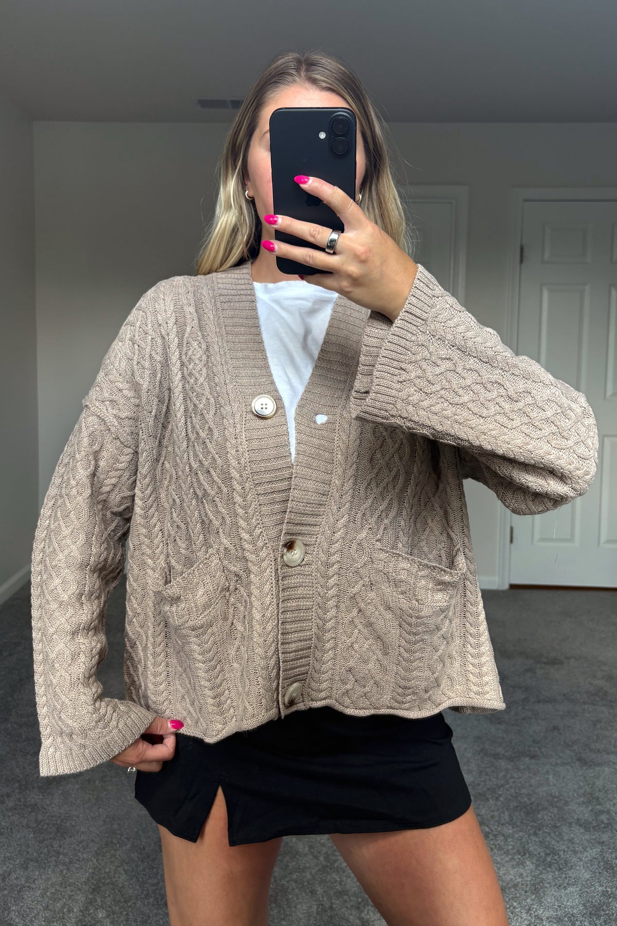 Fireside beige cable knit cardigan layered over a white top and black skirt, showing its V-neckline, large buttons, and front patch pockets.

