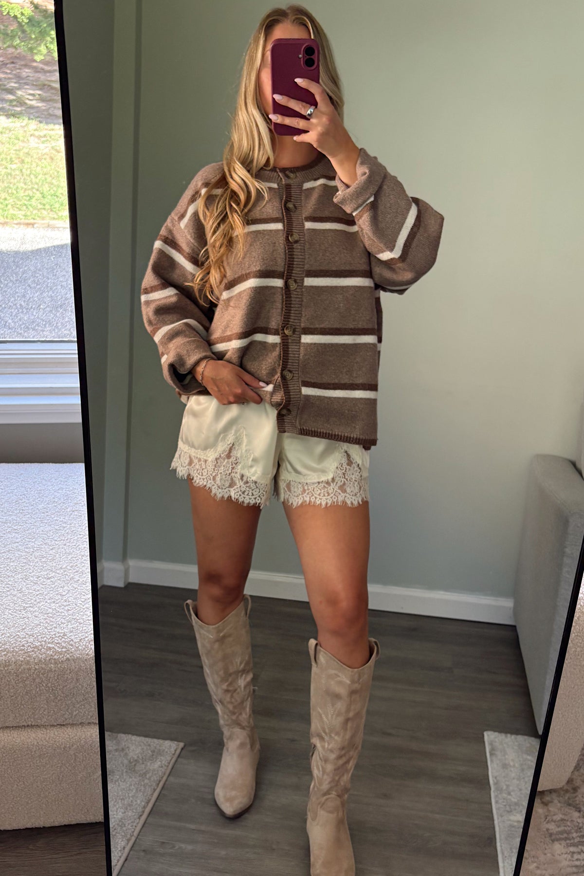A woman takes a mirror selfie in a room, wearing a brown and beige striped knit cardigan, cream lace-trimmed shorts, and beige knee-high boots.