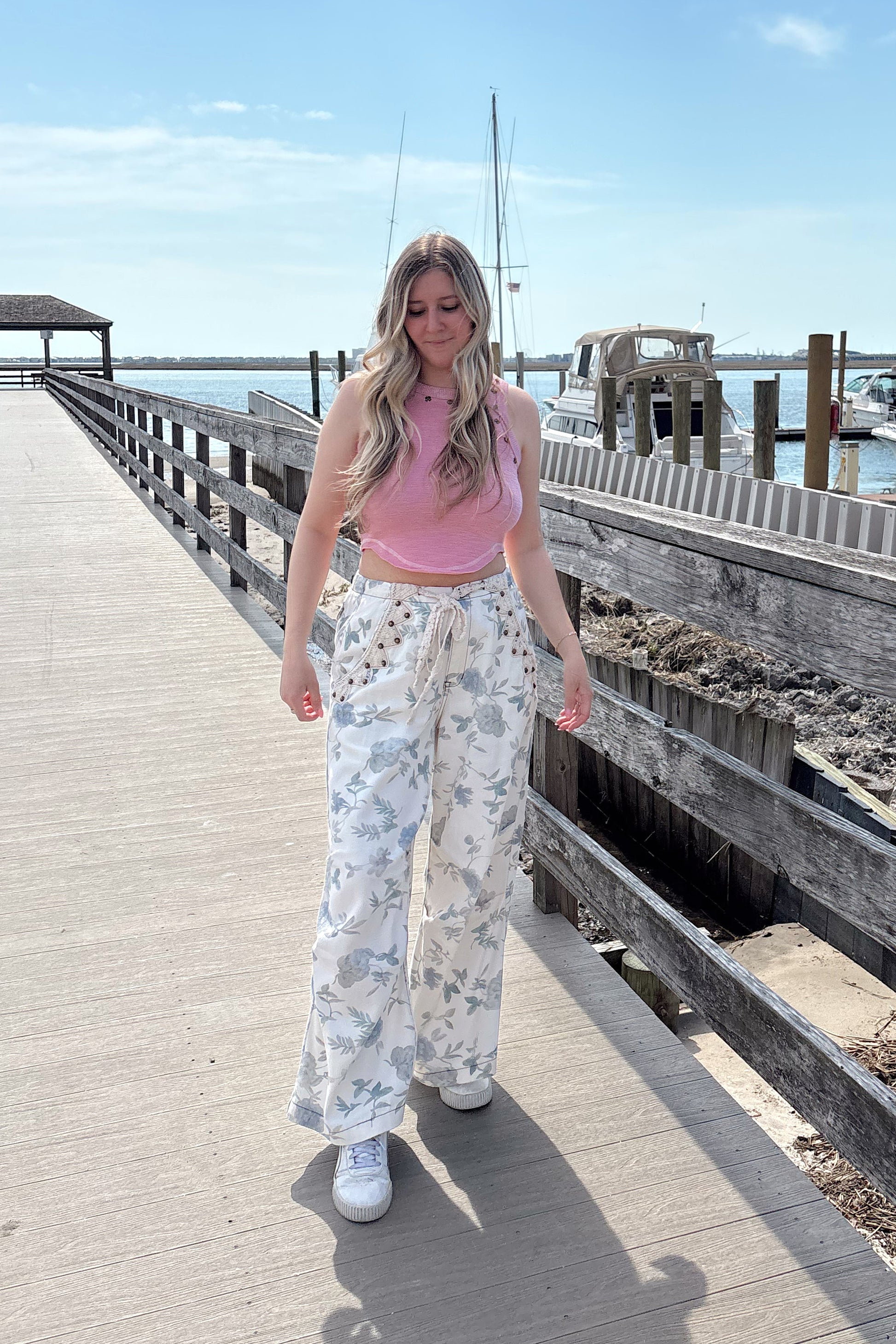 Woman in a pink top and floral pants standing on a wooden pier with a clear blue sky.