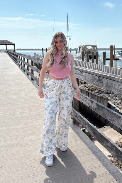 Woman in a pink top and floral pants standing on a wooden pier with a clear blue sky.