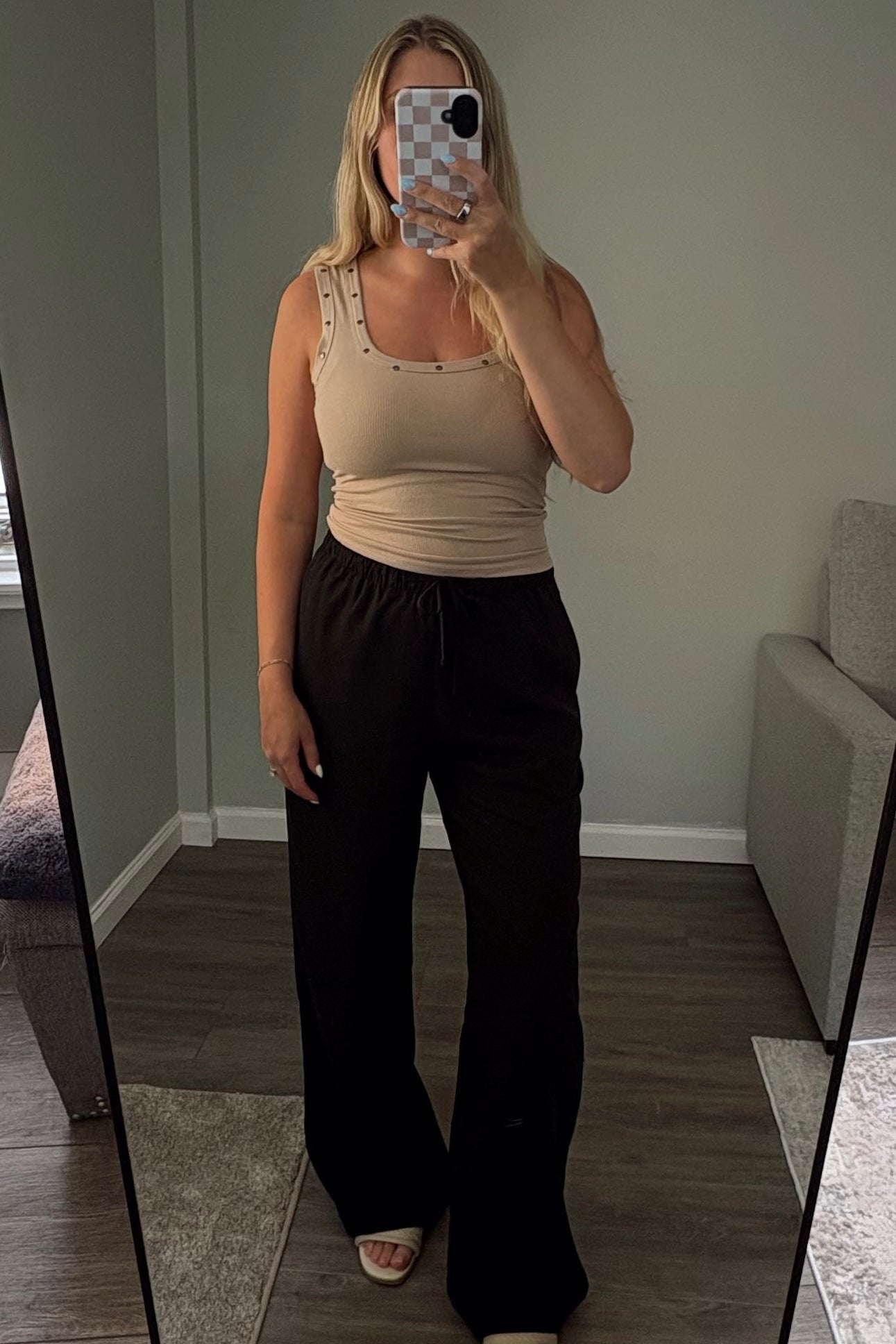 Person taking a mirror selfie in a room with a large mirror wearing black linen pants and a beige tank top