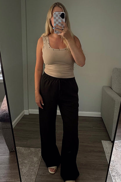 Person taking a mirror selfie in a room with a large mirror wearing black linen pants and a beige tank top