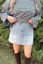 Person wearing a striped shirt, denim skirt, and brown knee-high boots standing in front of green bushes.