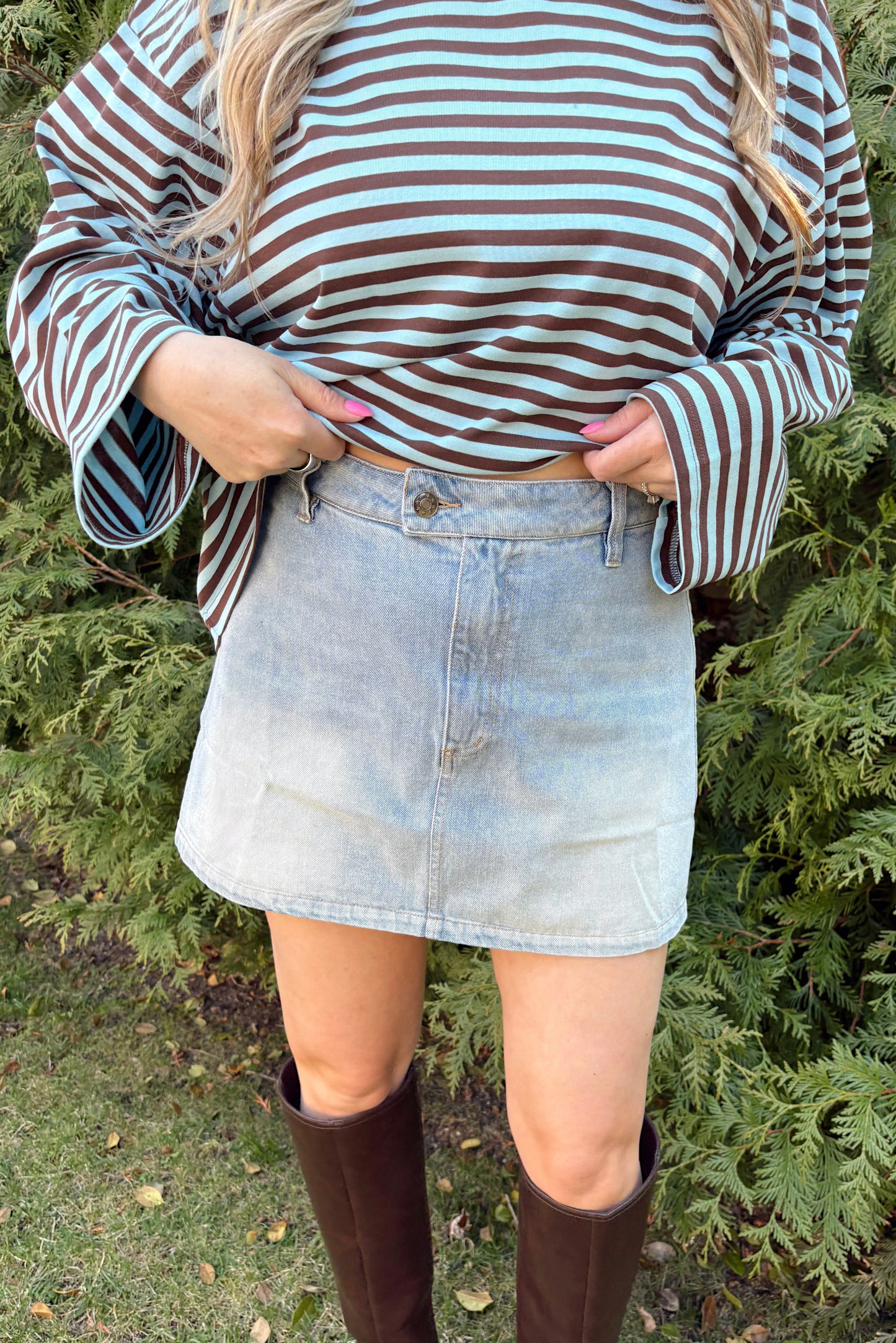 Person wearing a striped shirt, denim skirt, and brown knee-high boots standing in front of green bushes.