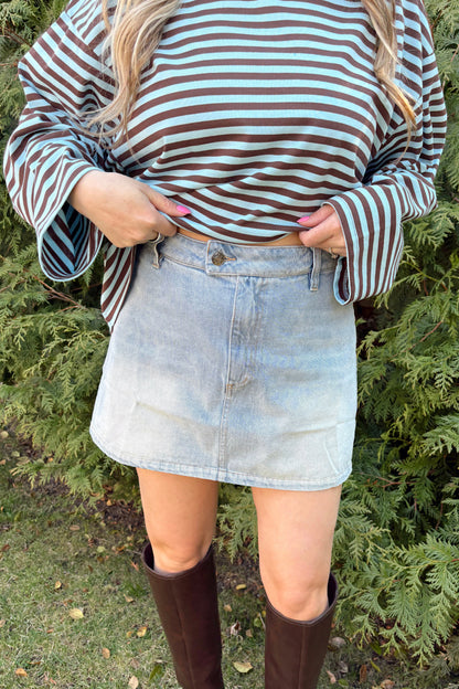 Person wearing a striped shirt, denim skirt, and brown knee-high boots standing in front of green bushes.