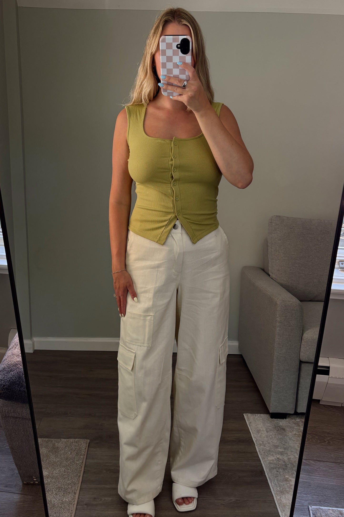 Woman taking a mirror selfie wearing a green top and white pants in a room with a couch and window.