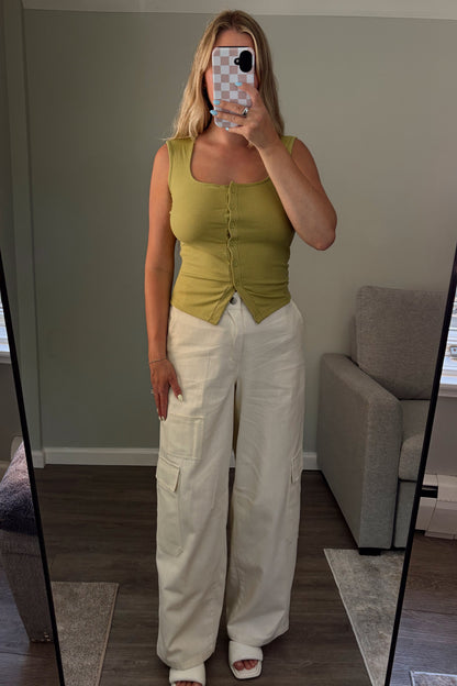 Woman taking a mirror selfie wearing a green top and white pants in a room with a couch and window.