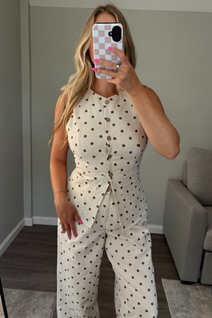 A woman takes a mirror selfie wearing a cream-colored sleeveless top with brown polka dots. The top features a button-up front, a fitted waist, and she is wearing matching wide-leg pants.