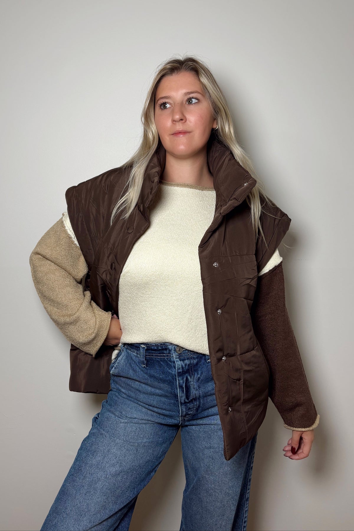 Parker brown puffer vest worn open over a cream and brown colorblock sweater with blue jeans, showing its high collar and extended shoulders.

