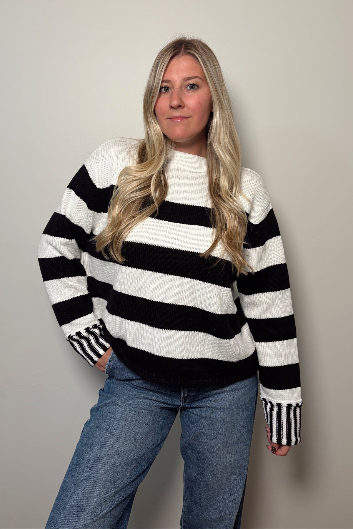 Front view of the Sunday Brunch black and white striped sweater worn untucked with blue jeans, showing its boxy fit and contrast striped cuffs.

