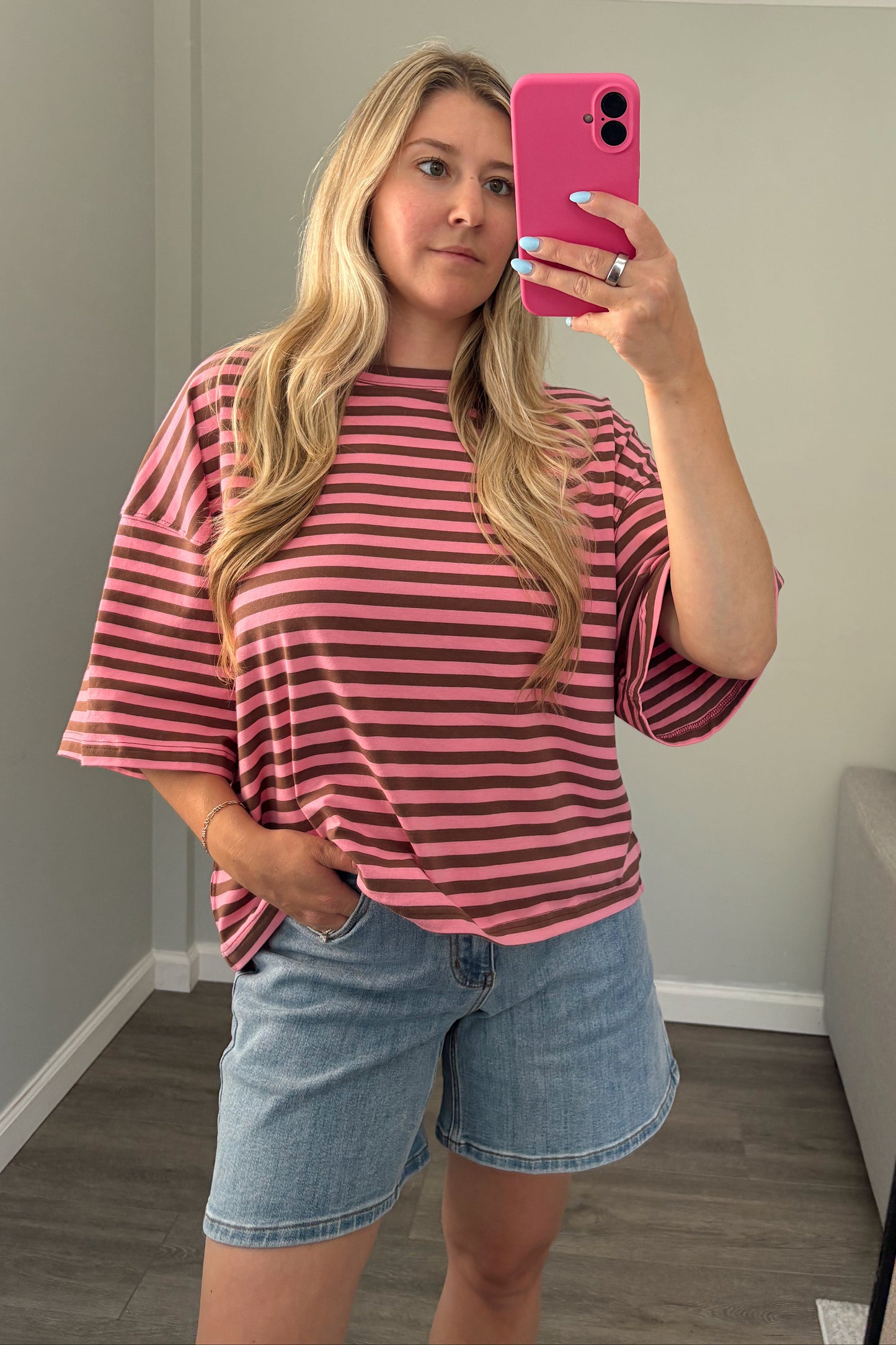 A woman takes a mirror selfie wearing a pink and dark brown striped short-sleeve t-shirt and light blue denim shorts in a home setting.