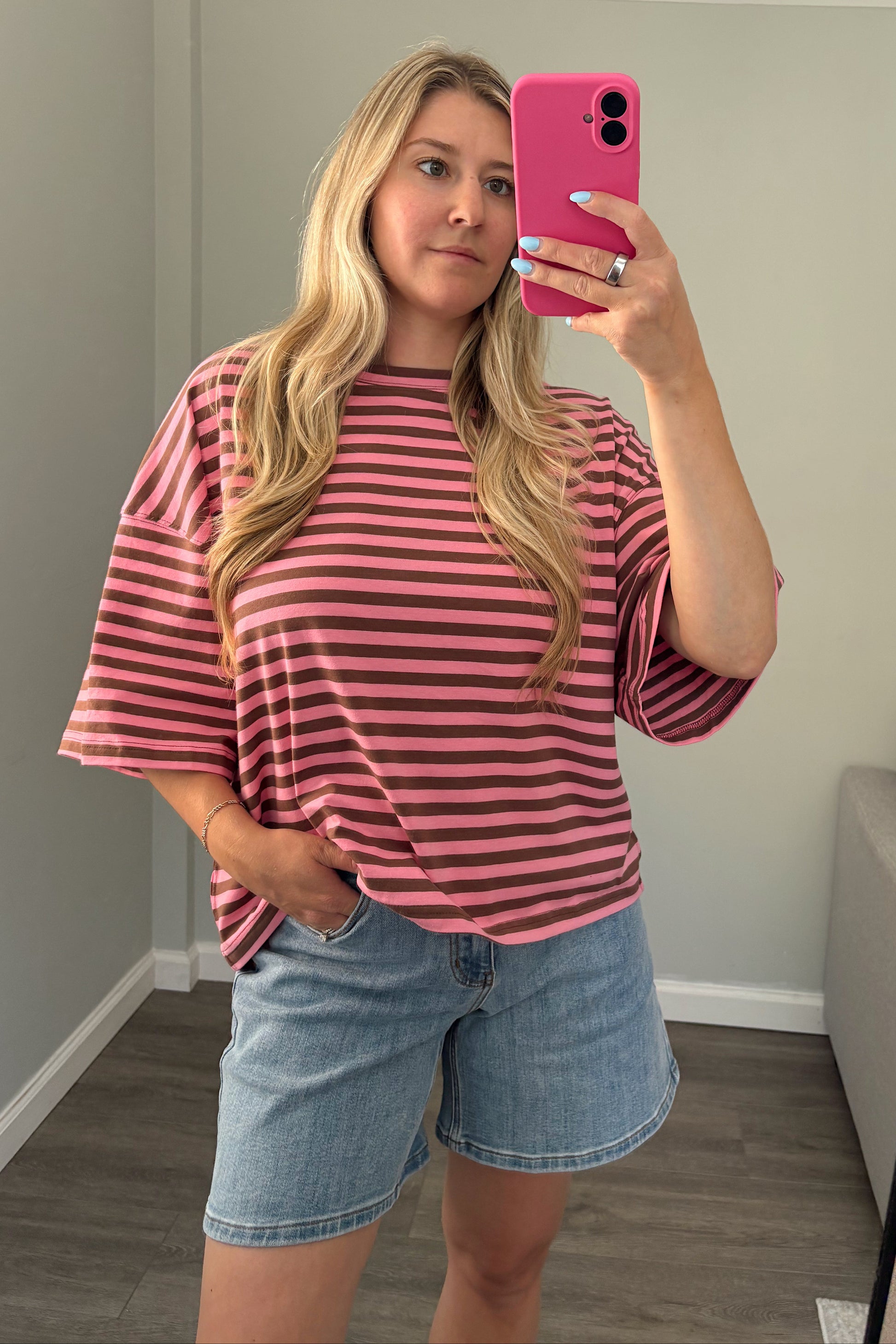 A woman takes a mirror selfie wearing a pink and dark brown striped short-sleeve t-shirt and light blue denim shorts in a home setting.