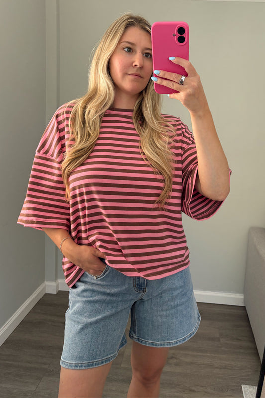 A woman takes a mirror selfie wearing a pink and dark brown striped short-sleeve t-shirt and light blue denim shorts in a home setting.