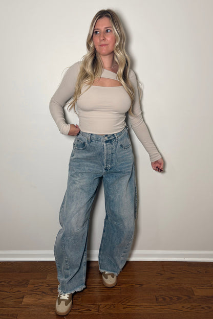 Woman wearing a beige top and blue jeans against a white wall.
