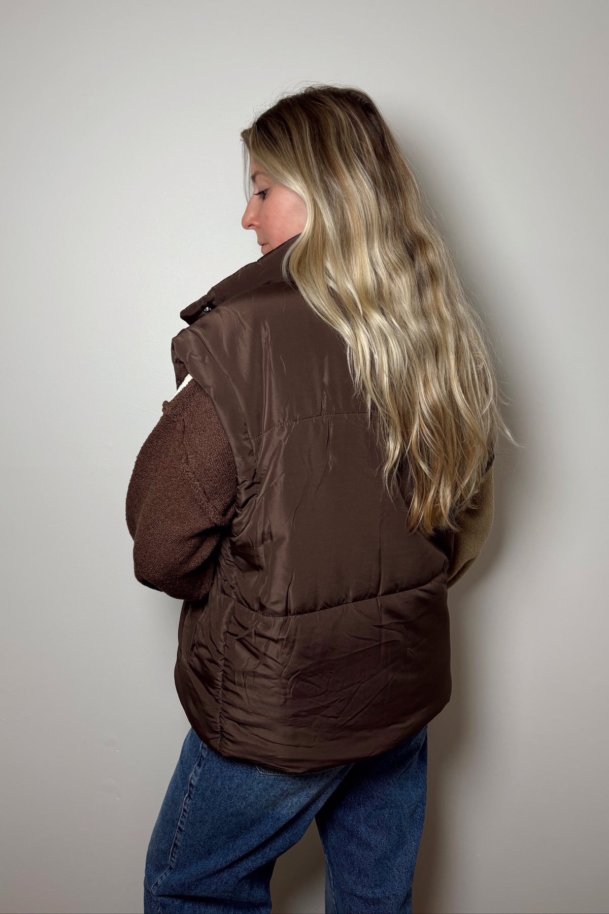 Back view of the Parker brown puffer vest showing its quilted panels, extended shoulders, and longer hem over blue jeans.

