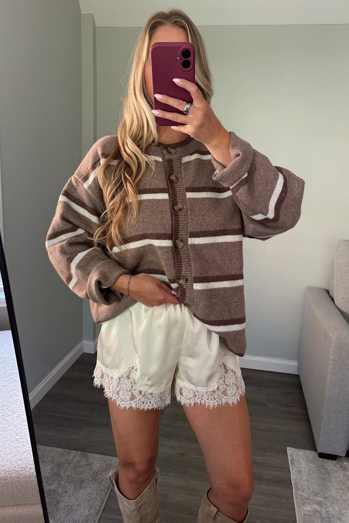 A woman takes a mirror selfie showing a brown and beige striped knit cardigan sweater, cream lace-trimmed shorts, and beige boots in a home setting.