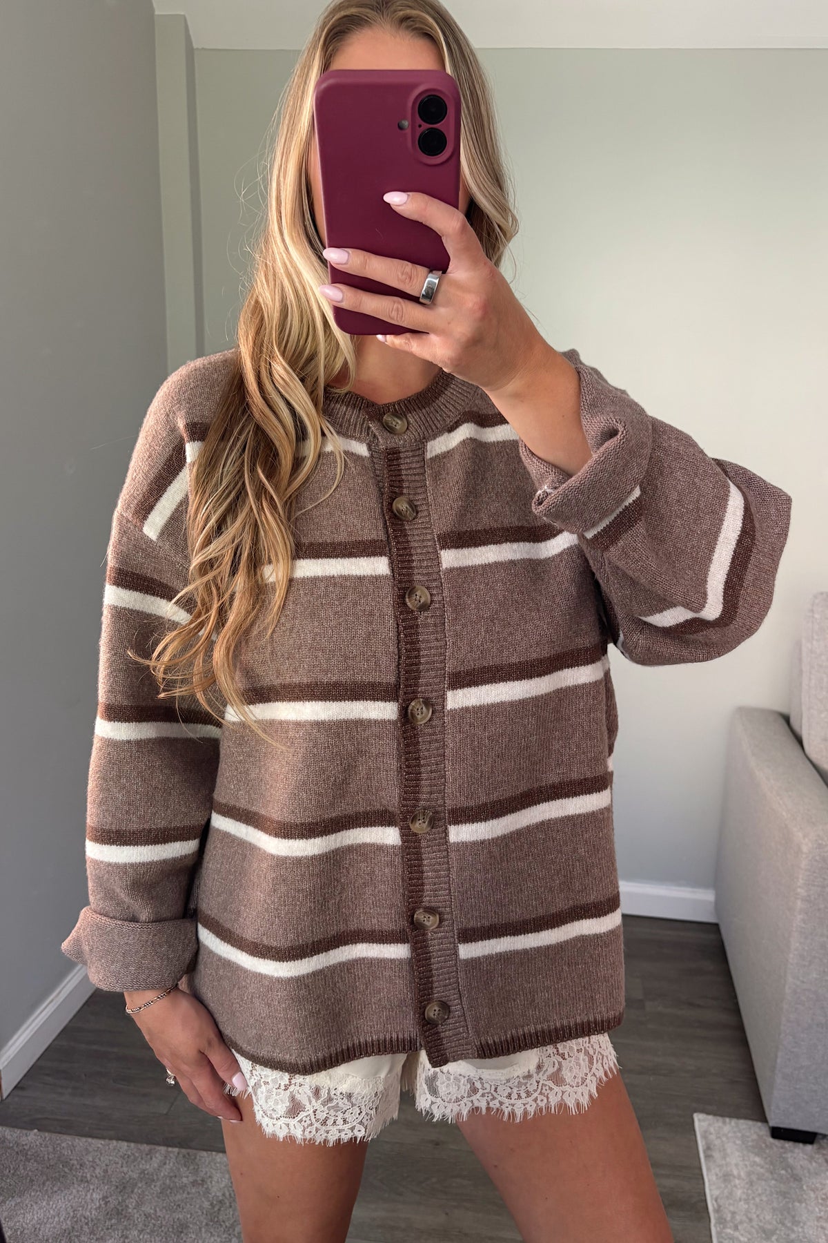 A woman takes a mirror selfie wearing a brown and cream striped knit cardigan with lace hem shorts in a living room setting.
