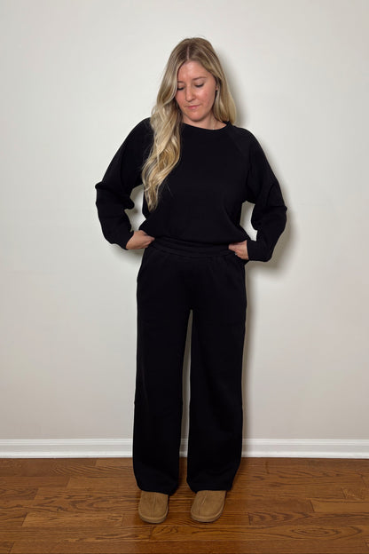 Woman wearing a black lounge set standing against a plain wall.