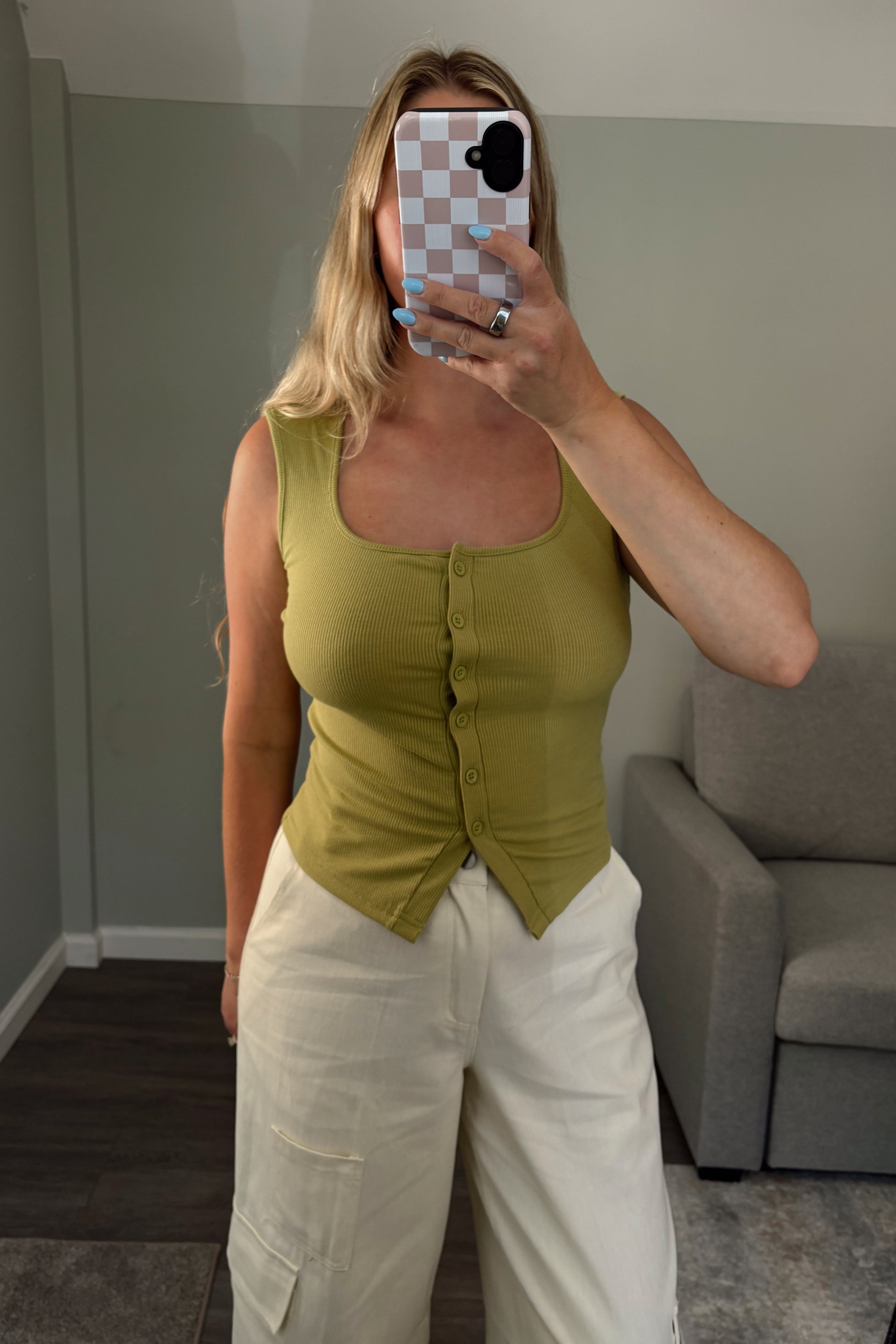 Woman taking a mirror selfie wearing a green sleeveless top and white pants in a room with a gray couch.