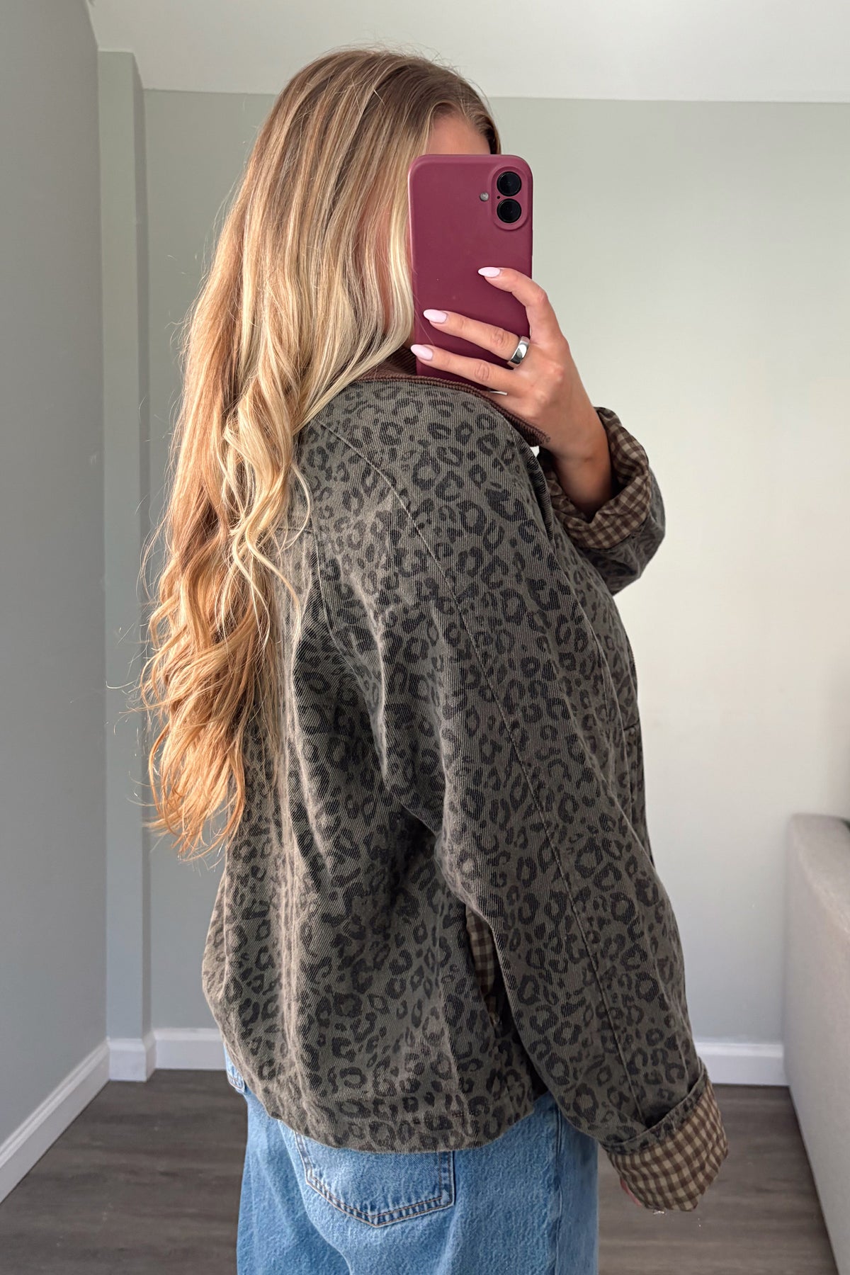 A woman takes a mirror selfie in a room, wearing an oversized dark olive green and brown leopard print cardigan with blue jeans.