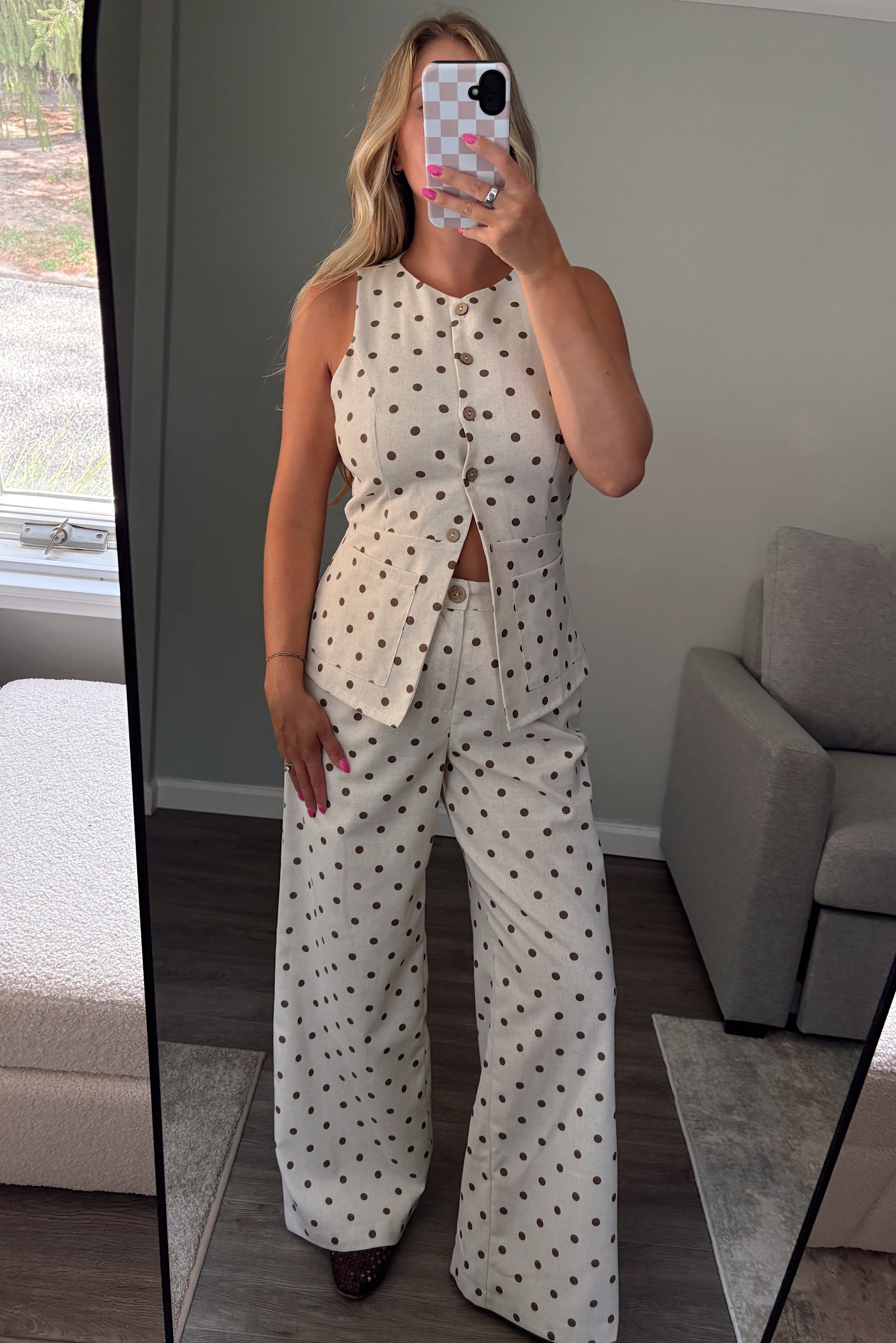 A woman takes a mirror selfie in a bedroom showing a matching two-piece outfit consisting of a sleeveless button-up top and wide-leg pants, both in an off-white fabric with a dark brown polka dot pattern.