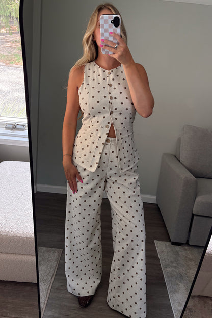 A woman takes a mirror selfie in a bedroom showing a matching two-piece outfit consisting of a sleeveless button-up top and wide-leg pants, both in an off-white fabric with a dark brown polka dot pattern.