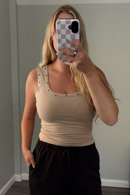 Woman taking a mirror selfie wearing a beige tank top and black pants in a room with a gray couch.