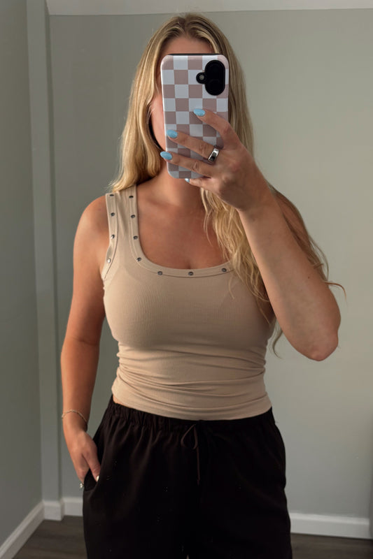 Woman taking a mirror selfie wearing a beige tank top and black pants in a room with a gray couch.