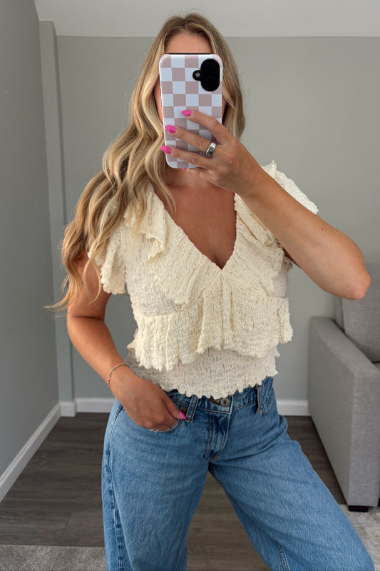 A woman takes a mirror selfie wearing a cream ruffled knit top and blue jeans in a room with gray walls and a gray sofa.