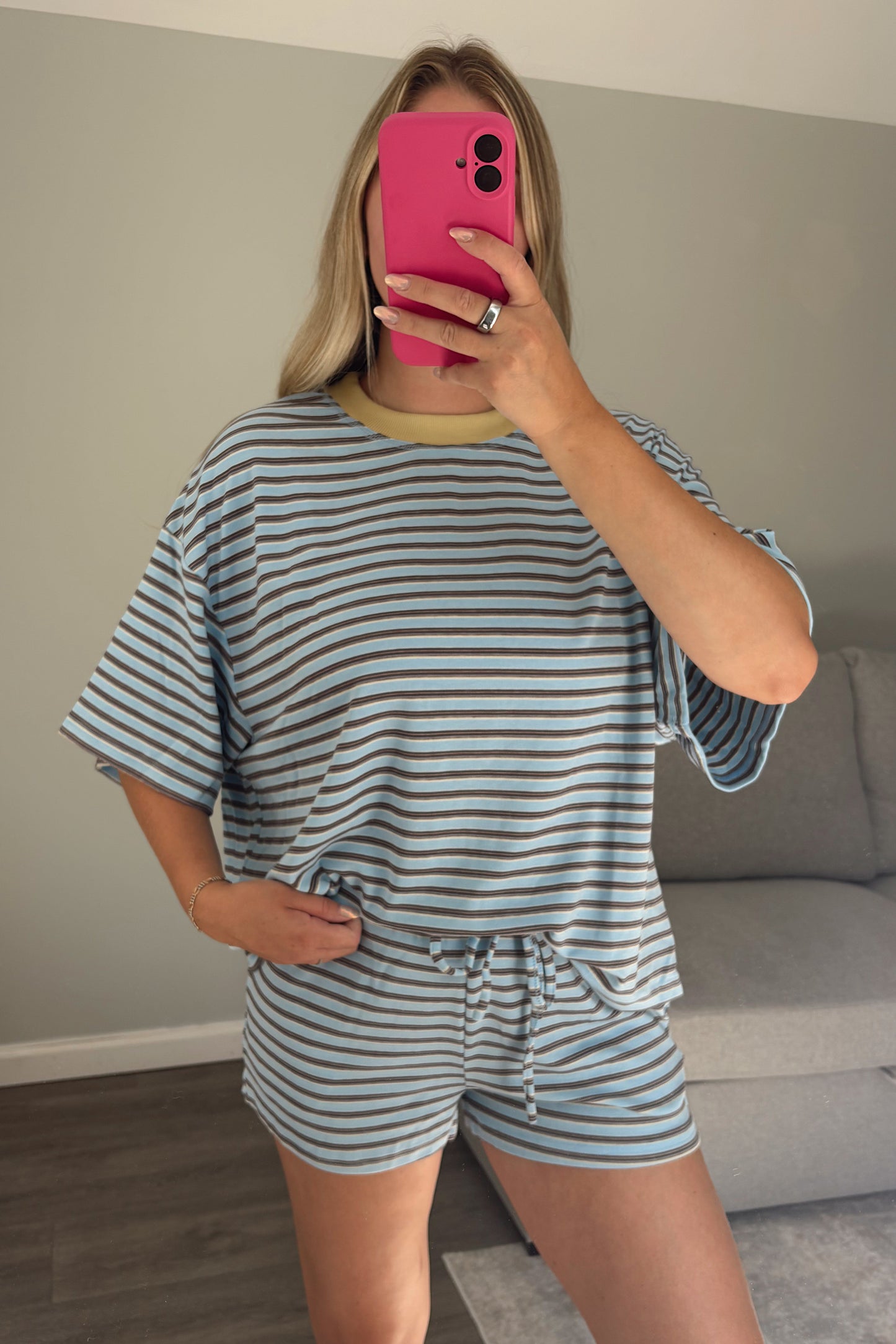 A woman takes a mirror selfie in a living room wearing a matching light blue and white striped lounge set consisting of a short-sleeve top and shorts with a drawstring waist.