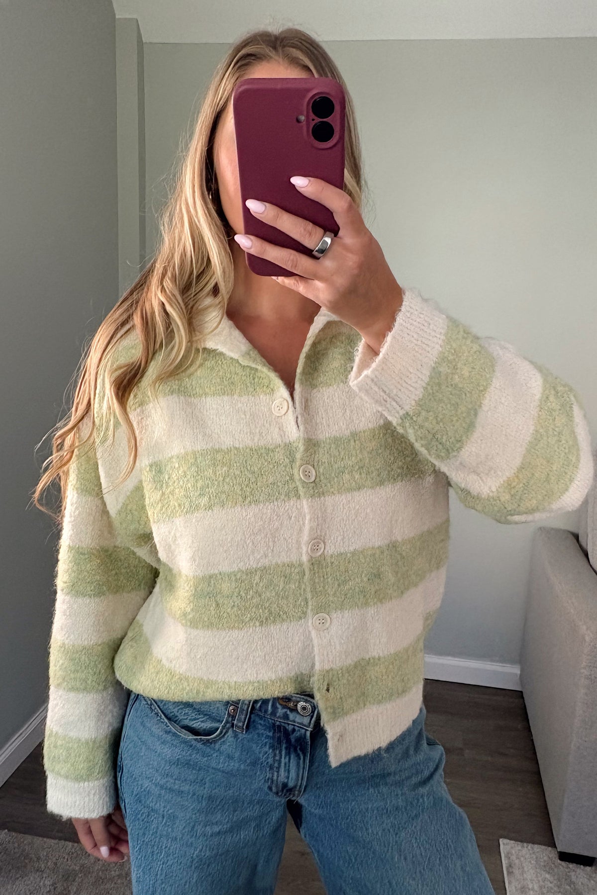 A woman takes a mirror selfie wearing a fuzzy, long-sleeve button-up cardigan with horizontal sage green and cream stripes, paired with blue jeans in a home setting.