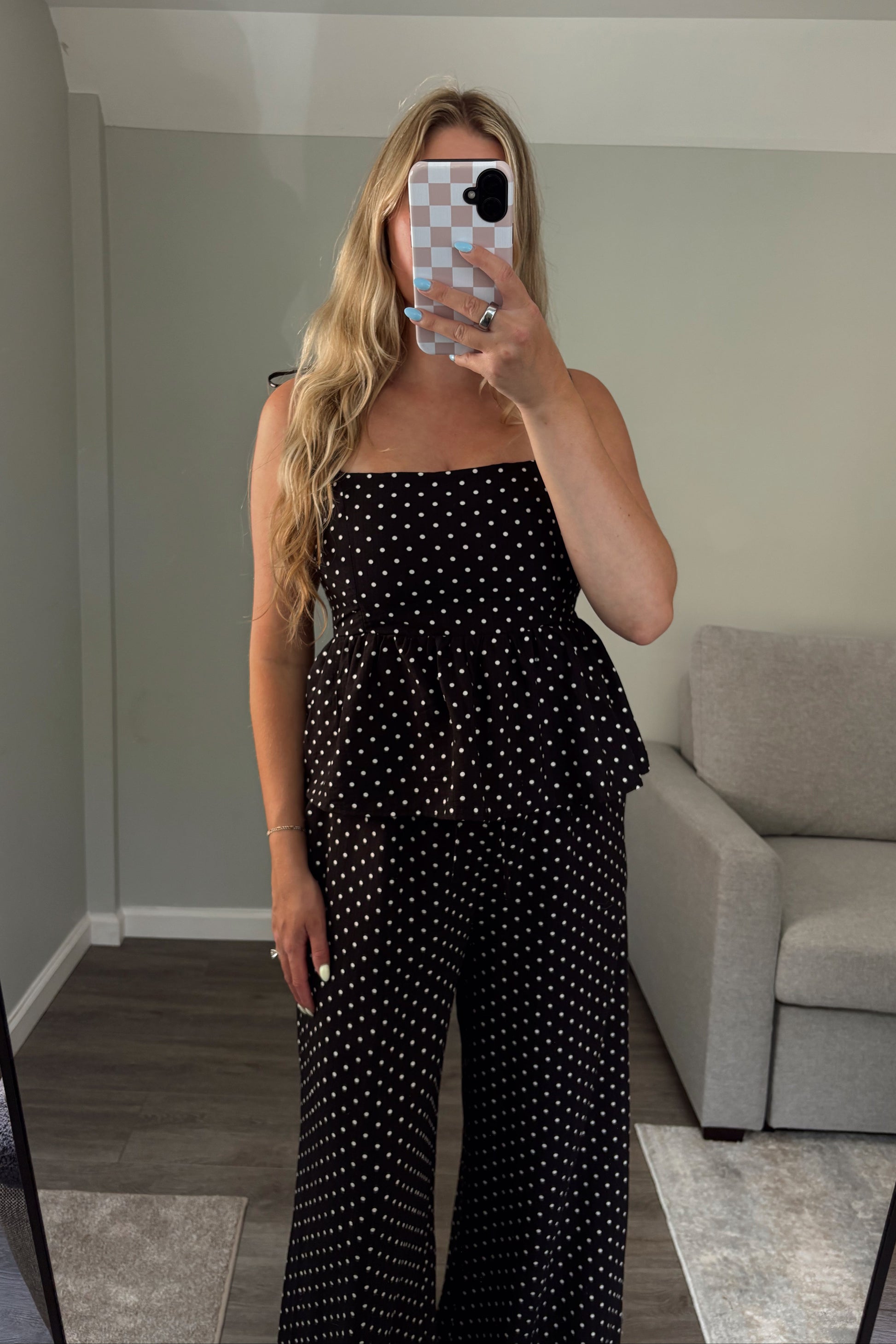 Woman taking a mirror selfie wearing a black polka dot outfit in a room with a gray couch.