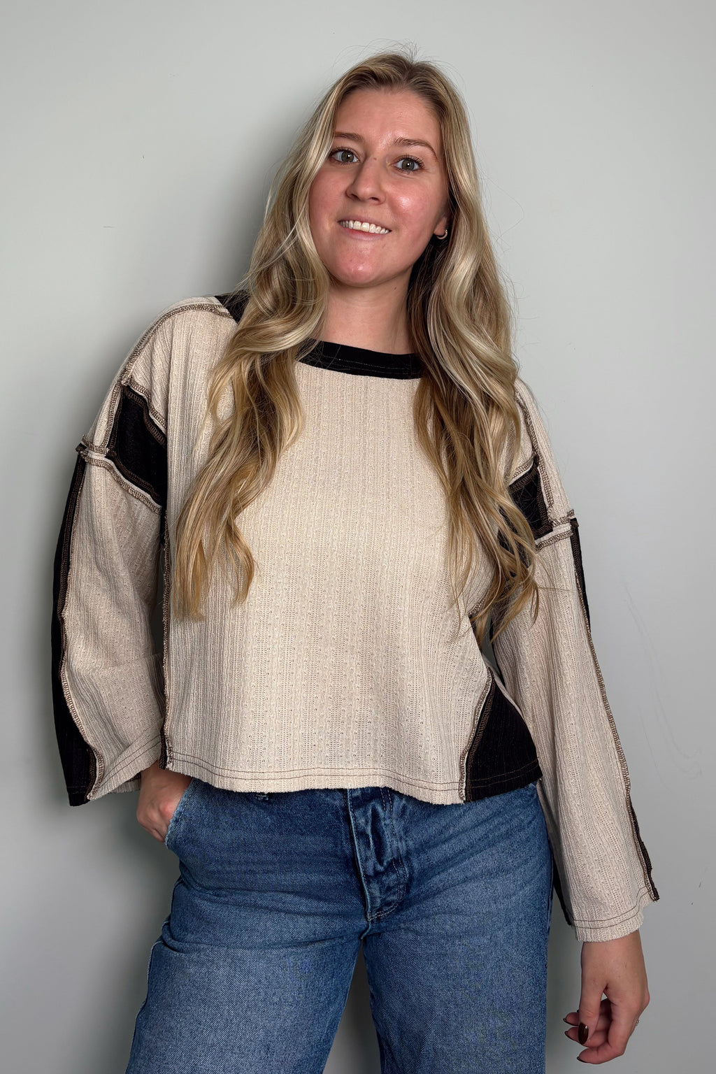 Smiling model wearing the Sawyer Colorblock Knit Top, a cream and black colorblock top with a boat neckline and exposed seams, styled with jeans