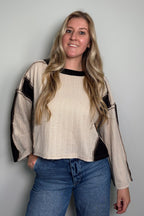 Smiling model wearing the Sawyer Colorblock Knit Top, a cream and black colorblock top with a boat neckline and exposed seams, styled with jeans
