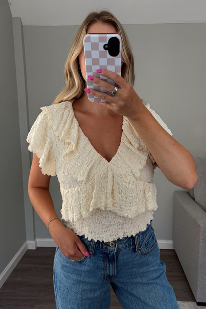 A woman takes a mirror selfie wearing a cream ruffled knit top and blue jeans in a room with gray walls and a gray sofa.