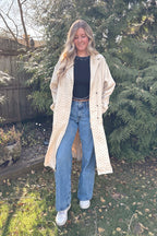 Woman wearing the beige Manhattan polka dot trench coat open over a black top and blue jeans outdoors