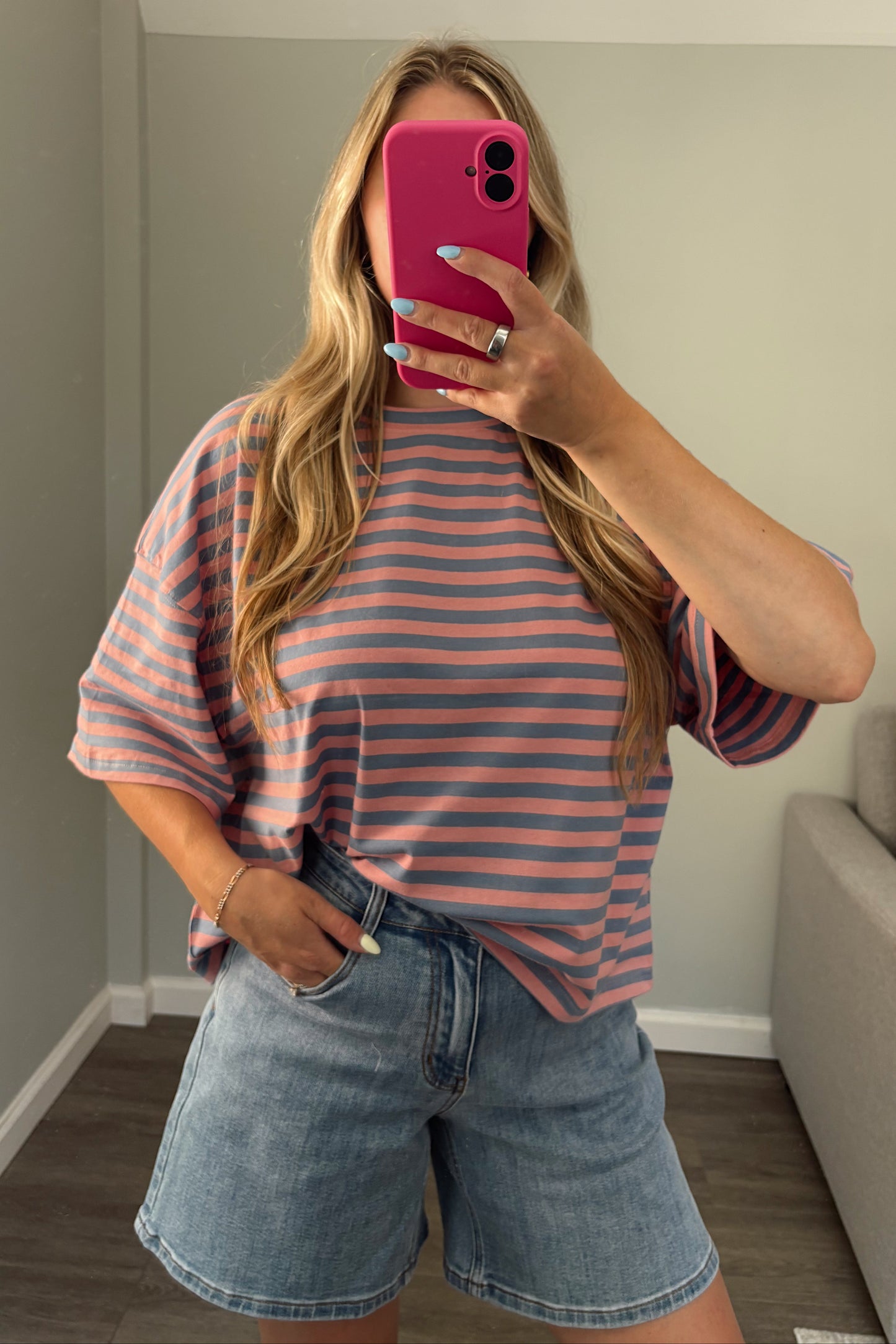 A woman takes a mirror selfie wearing a pink and blue horizontally striped t-shirt and light-wash denim shorts in a home setting.